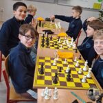 Sześciu uśmiechniętych chłopców gra w szachy w klasie, siedząc parami przy dwóch stolikach z szachownicami.