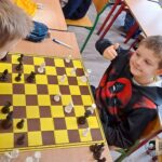 Dwóch chłopców gra w szachy przy stole; jeden z nich uśmiecha się i pokazuje kciuk w górę do kamery.