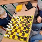 Dwoje dzieci siedzi przy stole i gra w szachy na żółto-brązowej szachownicy w klasie.
