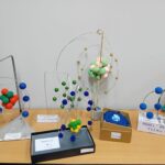 Modele atomów wykonane z kolorowych kulek i drutów wyświetlane na drewnianym stole na tle białej ściany.