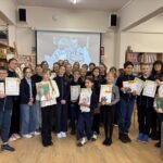 Grupa dzieci pozuje z certyfikatami i książkami w bibliotece, uśmiechając się do kamery.