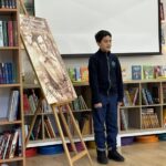 Chłopiec stoi obok dużego portretu i półek z książkami w klasie lub bibliotece.