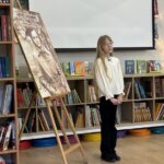 Dziewczyna stoi przy portrecie na sztaludze w bibliotece, za nią znajdują się półki z książkami i projektor.