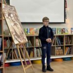 Chłopiec stoi przy portrecie na sztaludze w bibliotece, z książkami i ekranem projekcyjnym za nim.