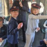 Dwoje dzieci w czapkach konduktorskich trzyma bilety w tramwaju, a dorosły konduktor przemawia za nimi.