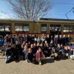 Duża grupa dzieci pozuje i uśmiecha się przed nieruchomym żółtym pociągiem w słoneczny dzień.