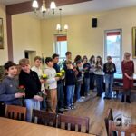 Grupa uczniów i nauczyciel stoją w pokoju, każdy z nich trzyma małą roślinę doniczkową.