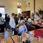 Grupa studentów wręcza kwiaty starszym osobom siedzącym przy stolikach w jasnym, wesołym pomieszczeniu.