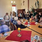 Grupa dzieci i osób starszych dzieli się jedzeniem i rozmawia przy stole w jasnym pomieszczeniu.