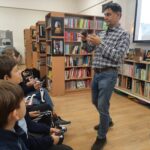 Mężczyzna przemawia do siedzących w bibliotece dzieci, trzymając jakiś przedmiot, podczas gdy one patrzą i słuchają.