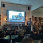 Mężczyzna przedstawia prezentację na temat statku uczniom siedzącym w klasie przypominającej bibliotekę.