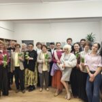 Grupa seniorów i nastolatków pozujących razem w pomieszczeniu, niektórzy trzymają kwiaty, w otoczeniu klasy.