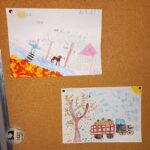 Dwa jesienne rysunki dzieci są przypięte do tablicy korkowej, przedstawiając drzewa, liście i ciężarówkę.