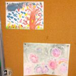 Dwie dziecięce akwarele przypięte do tablicy korkowej, przedstawiające drzewa i abstrakcyjne kształty.