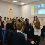 Dzieci stoją i podnoszą ręce w klasie, stojąc przed prezentacją wyświetlaną na ekranie.