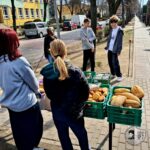 Pięć osób stoi na chodniku przy stolikach z koszami chleba i jedzenia, niedaleko ulicy.