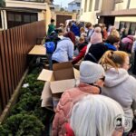 Grupa ludzi zbiera się na zewnątrz przy stołach i kartonowych pudłach obok brązowego drewnianego płotu.