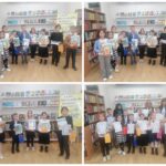 Cztery grupy dzieci trzymające certyfikaty i książki, stojące w bibliotece z regałami.