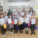 Dzieci trzymające certyfikaty i torby z prezentami pozują z dorosłymi w bibliotece, uśmiechając się do grupowego zdjęcia.