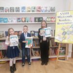 Troje dzieci z certyfikatami stoi w bibliotece obok żółtego znaku konkursu recytatorskiego.