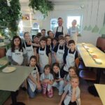 Grupa dzieci i dorosły w fartuchach pozują razem w jasnej, wypełnionej roślinami restauracji.