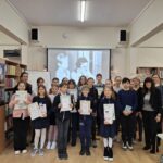 Grupa dzieci i dorosłych pozuje w bibliotece, trzymając certyfikaty, a za nimi wyświetlane jest czarno-białe zdjęcie.