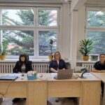 Trzy kobiety siedzą przy biurkach z laptopami i papierami w jasnym biurze z dużymi oknami i roślinami.