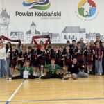 Młodzieżowa drużyna sportowa świętuje z medalami i szalikami na parkiecie sali gimnastycznej, z trenerami i kibicami.