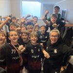 Młodzieżowa drużyna sportowa i trener świętują razem, uśmiechając się i unosząc pięści w szatni.