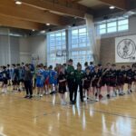 Drużyny piłki ręcznej chłopców stoją w szeregu na krytym boisku, przygotowując się do meczu lub wydarzenia.