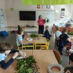 Nauczyciel pokazuje rośliny dzieciom siedzącym przy biurkach z materiałami rzemieślniczymi w jasnej klasie.