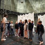 Grupa dzieci stoi na scenie teatralnej, słuchając dorosłego przed udrapowanym tłem.