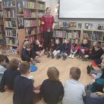 Kobieta przemawia do grupy dzieci siedzących w kręgu na podłodze w bibliotece.