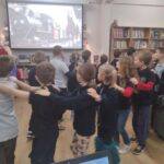 Dzieci stoją w szeregu z rękami na ramionach, oglądając animowany pociąg na ekranie w bibliotece.