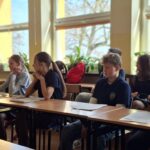 Pięciu uczniów siedzi przy biurkach w klasie w pobliżu okien, skupiając się na leżących przed nimi dokumentach.