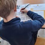 Dziecko o jasnobrązowych włosach rozwiązuje łamigłówki ołówkiem przy biurku w klasie.