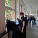 Uczniowie na szkolnym korytarzu, niektórzy patrzą przez okna, inni stoją i rozmawiają w grupach.