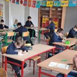 Dzieci w klasie siedzą przy biurkach, pisząc lub myśląc, z kolorowymi dziełami sztuki na ścianach.