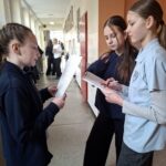 Trzy dziewczyny stoją na szkolnym korytarzu i czytają z gazet; w tle widać innych uczniów.