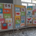 Odciski dłoni dzieci wyświetlane na trzech tablicach w jasnym pomieszczeniu z oknami w tle.