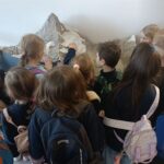 Grupa dzieci z plecakami przygląda się i dotyka dużego modelu góry w pomieszczeniu.