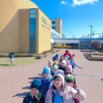 Grupa dzieci w kolorowych czapkach stoi w szeregu przed nowoczesnym budynkiem szkoły z cegły.