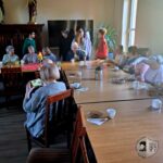 Grupa starszych osób i młodych dorosłych siedzi i stoi wokół stołu z kubkami i przekąskami.