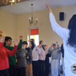 Grupa uczniów podnosi ręce, podczas gdy kobieta prowadzi ich w klasie.