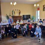 Grupa dzieci i osób starszych pozuje razem w jasnym pomieszczeniu ze stołami i dekoracjami.