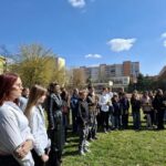 Grupa ludzi stoi na zewnątrz na trawie z budynkami i drzewami w tle pod błękitnym niebem.