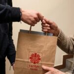 Dwie osoby wymieniają papierową torbę Caritas Poznań w pomieszczeniu, widoczne ręce, bez twarzy.