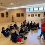 Grupa małych dzieci siedzi na podłodze galerii, słuchając przewodnika w muzeum sztuki.