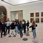 Dzieci gromadzą się wokół przewodnika w muzeum sztuki, z obrazami wyświetlanymi na białych ścianach.