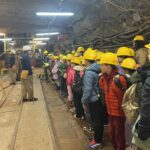 Dzieci w żółtych kaskach stoją w kolejce, słuchając przewodników wewnątrz podziemnego tunelu lub kopalni.